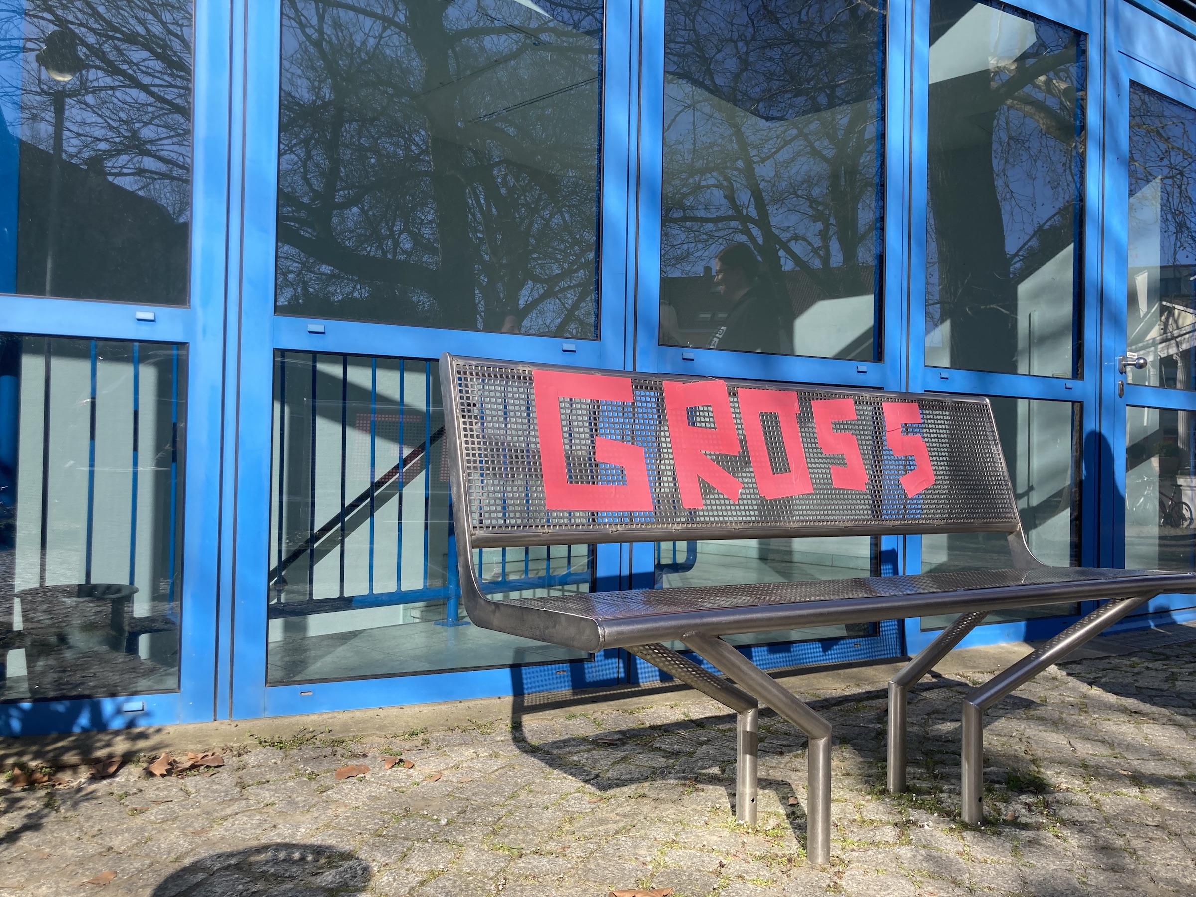 Projektbild im Freien. Zu sehen ist eine graue Bank, auf die mit rotem Klebeband das Wort „Gross“ geschrieben wurde. Im Hintergrund eine Fensterwand mit leuchtend blauen Rahmen.